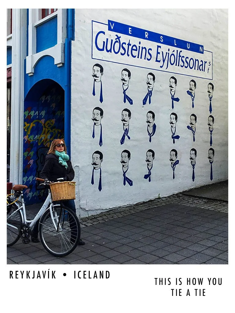 Woman with sunglasses and a scarf standing next to a bicycle with a basket, in front of a mural wall with portraits of a man with a mustache and tie, and a sign reading 'Verlsun Guðsteins Eyjólfsson r' in Reykjavik, Iceland.