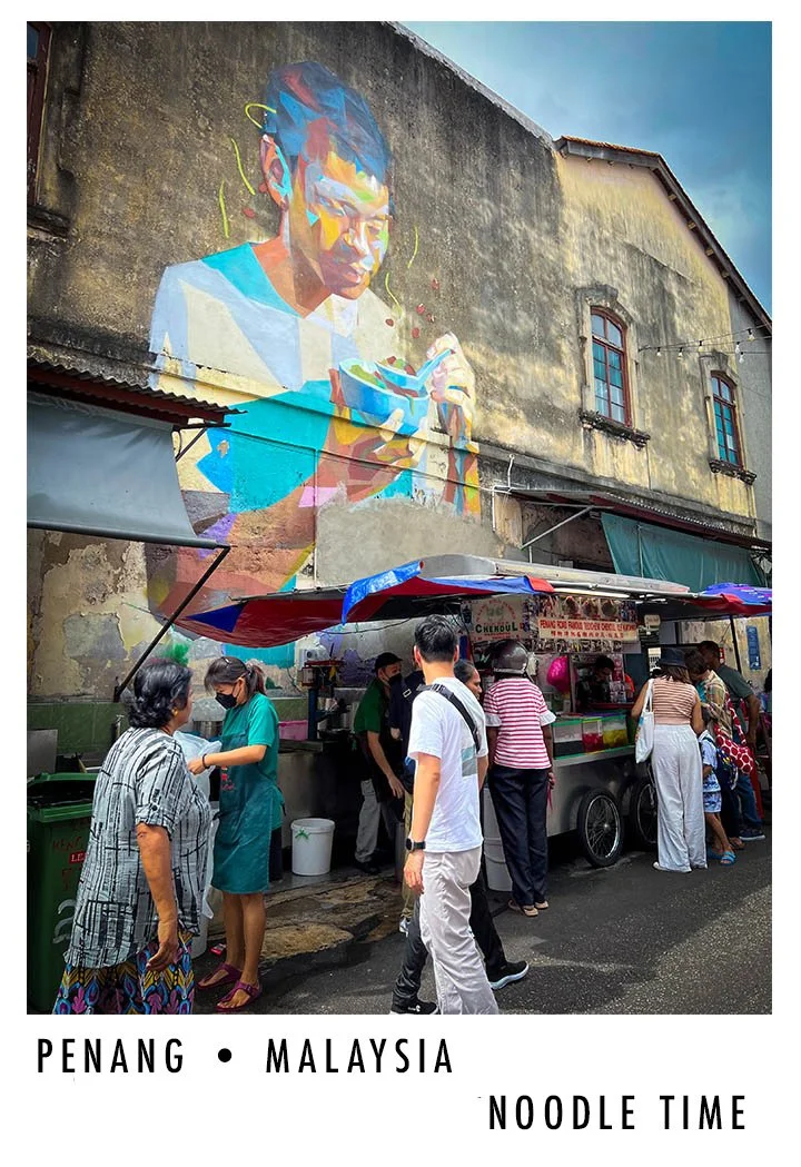 A street scene in Penang, Malaysia, featuring a colorful mural of a girl eating noodles on a weathered building wall. People are standing in line at a street food stall beneath the mural, with some engaging with vendors and others waiting.