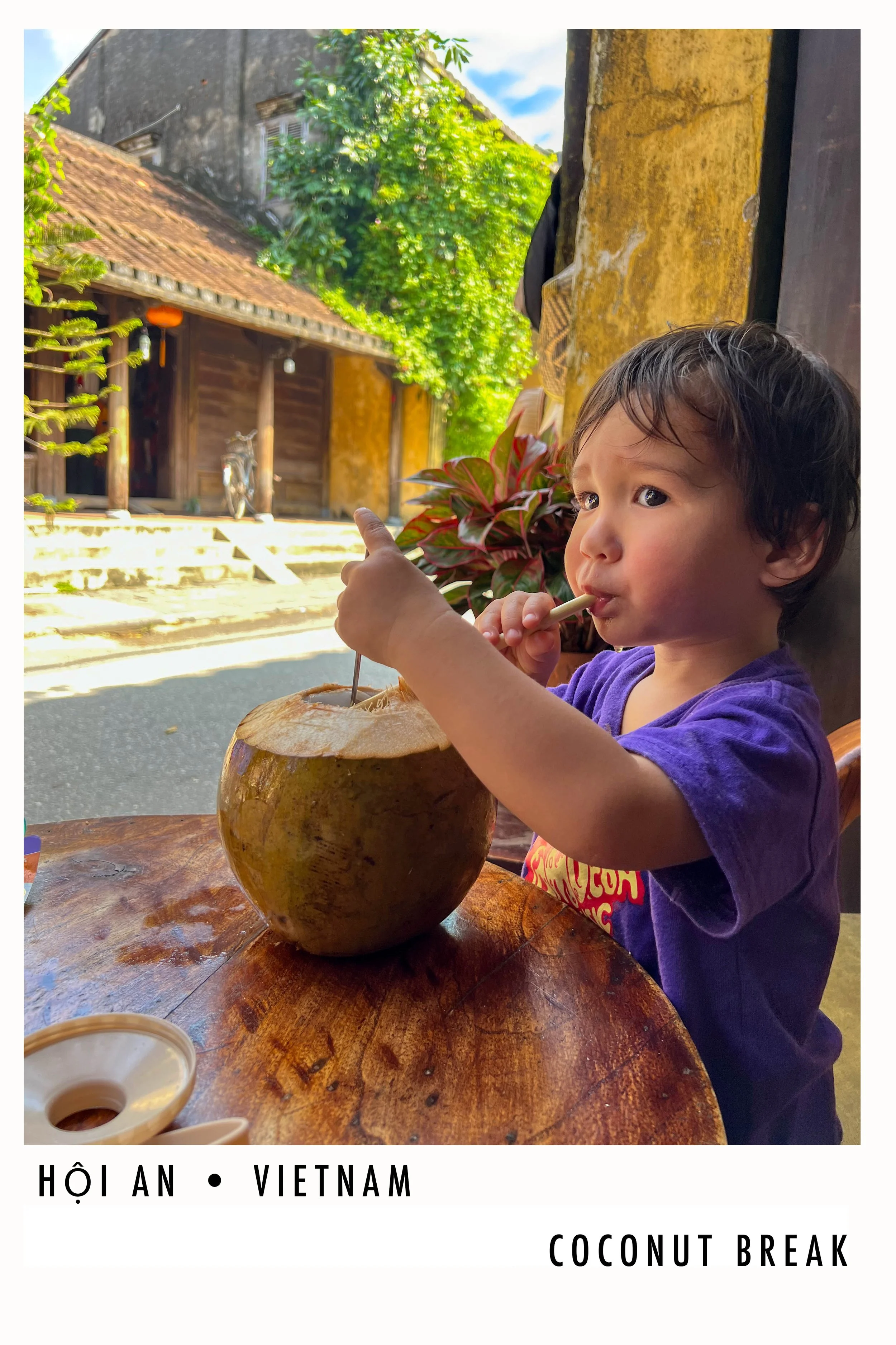 A young boy with wet hair sitting at a wooden table, sipping from a straw in a coconut. The coconut is on the table, and there is a potted plant with red and green leaves in the background. The scene appears to be outdoors with a rustic building and 