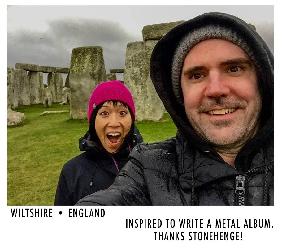A cheerful couple taking a selfie in front of Stonehenge on a cloudy day, both wearing jackets and head coverings, with the woman showing excitement and the man smiling.