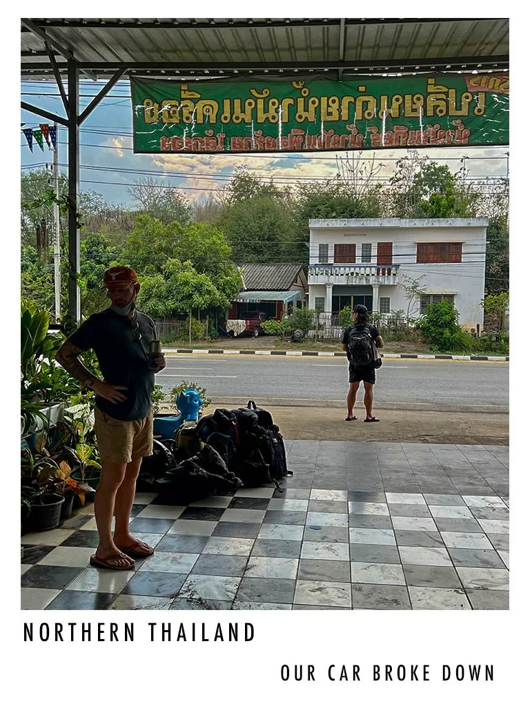 A person stands on a checkered tile floor with luggage and bottles, looking at a street with two people and white houses in northern Thailand. There is a large green sign with yellow and red script hanging above. The scene shows a cloudy sky and tree
