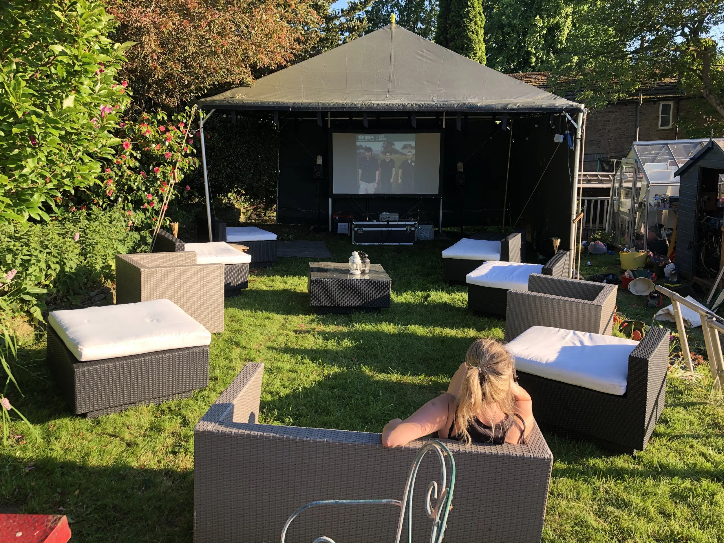 A backyard outdoor cinema setup with a large screen under a canopy, surrounded by outdoor furniture and a woman watching a movie on a grassy lawn in the evening.