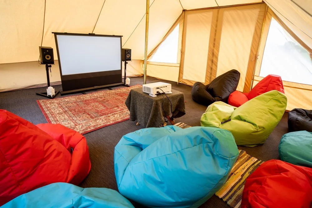 Tent interior with colorful bean bag chairs, a projector, speakers, and a film screen on a small table.
