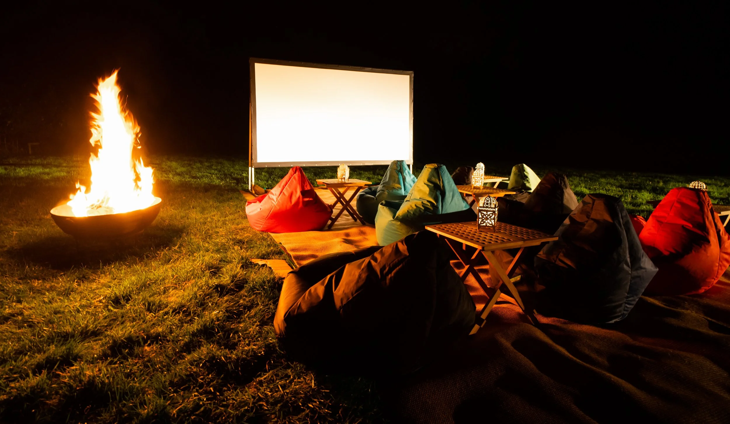 Outdoor movie night setup with bean bag chairs, small tables, lanterns, a large blank screen, and a fire pit on a grassy field at night.