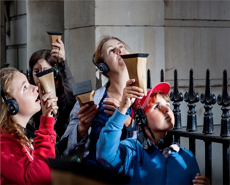 A group of people, including children and adults, standing outside near a black iron fence, holding and looking through listening devices called ear trumpets.