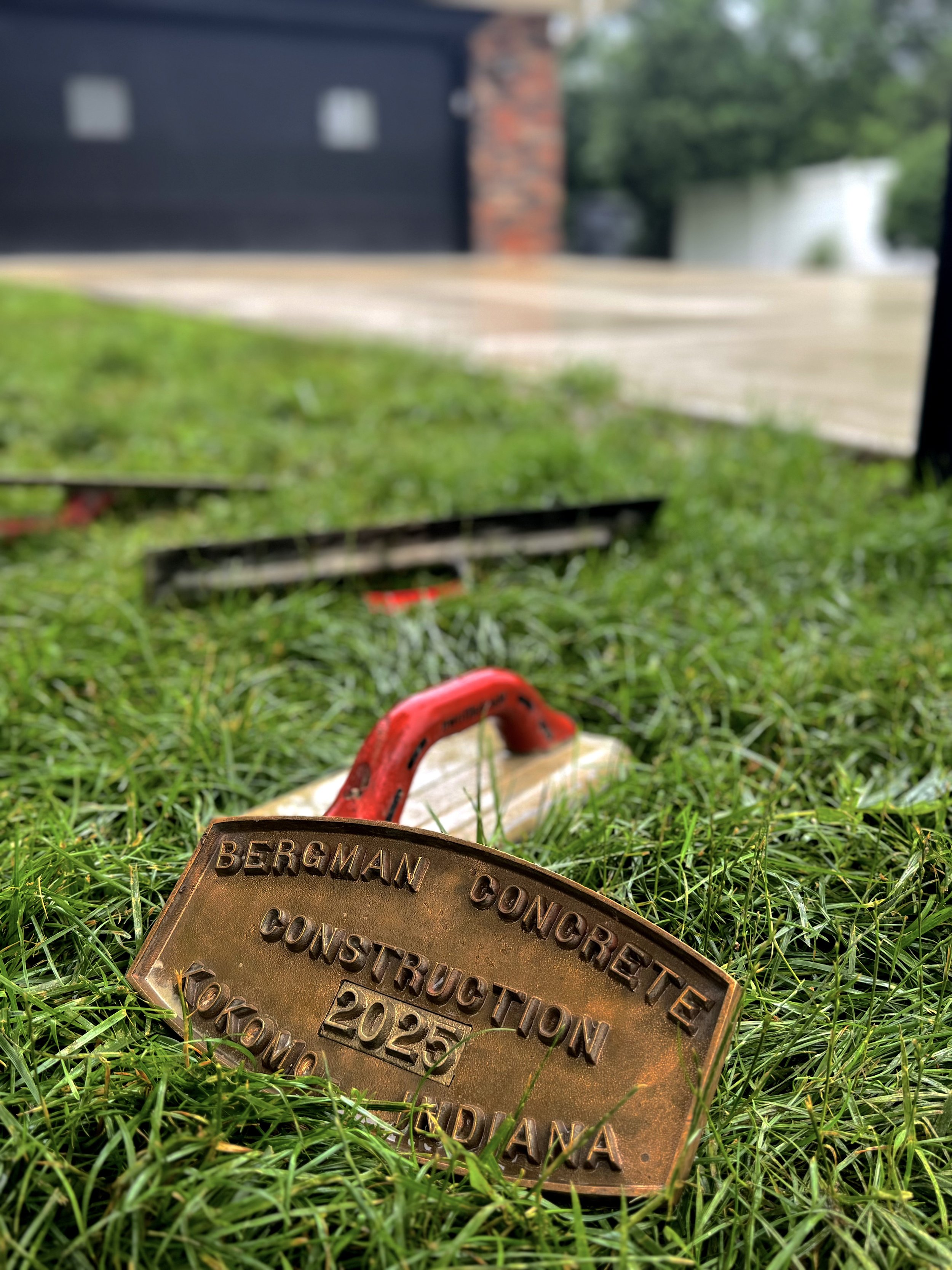 Bergman Concrete 2025 Stamp sitting in the grass with other concrete tools and a new concrete driveway in the background