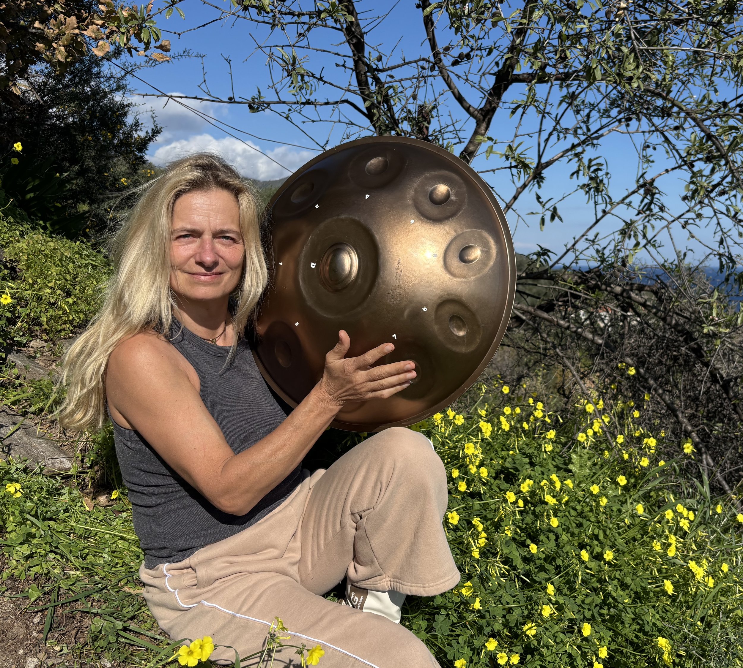 A woman with long blonde hair sitting outdoors among yellow flowers, holding a large handpan instrument, with trees and blue sky in the background.