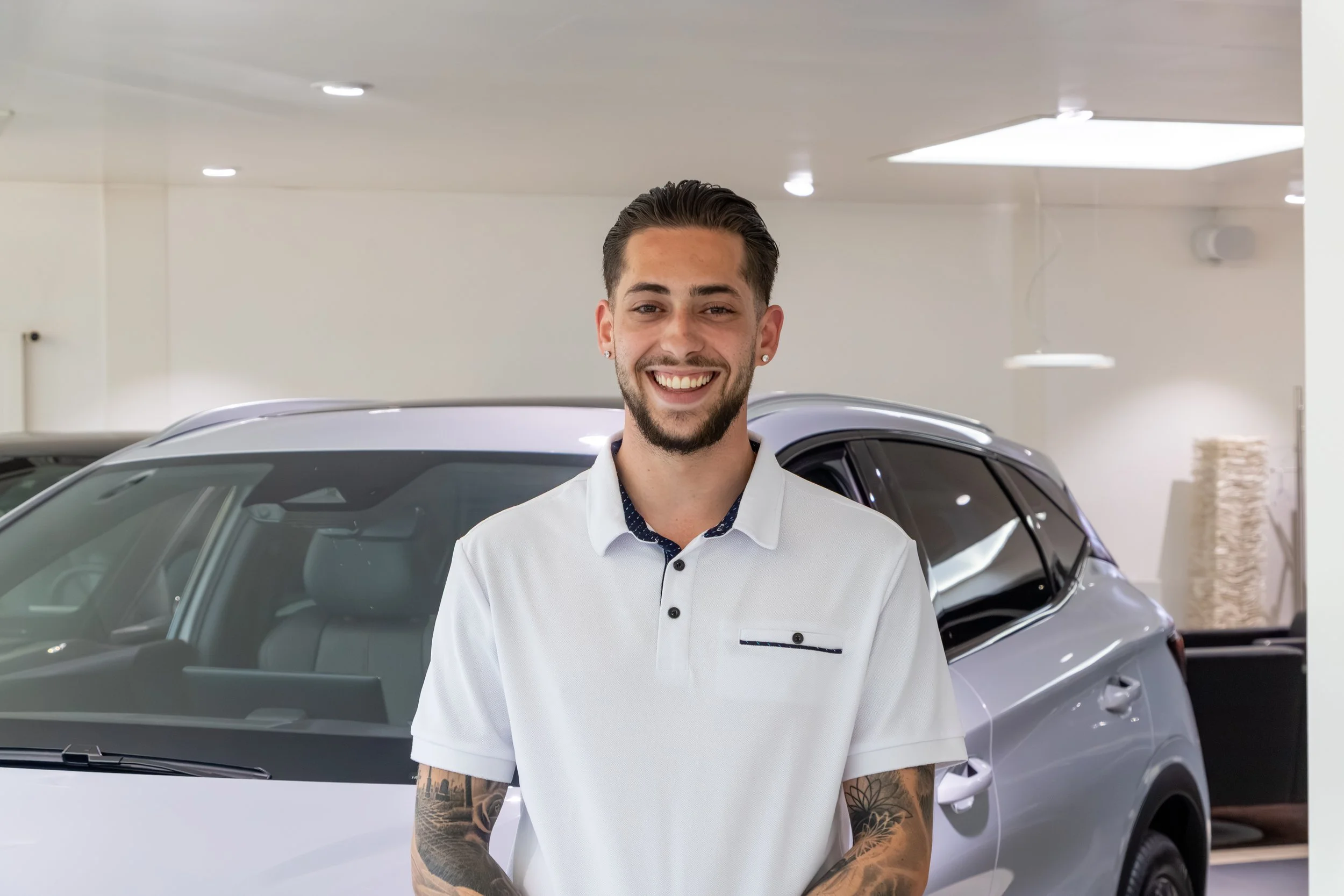 Ein lachender junger Mann mit Tattoos auf den Armen, trägt ein weißes Hemd und steht vor einem schwarzen Auto in einem hellen, modernen Raum.