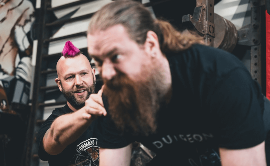 Two men with beards and black t-shirts in a gym, one with a pink mohawk and the other with long brown hair and a full beard, engaged in a log press. Strongman training in London.