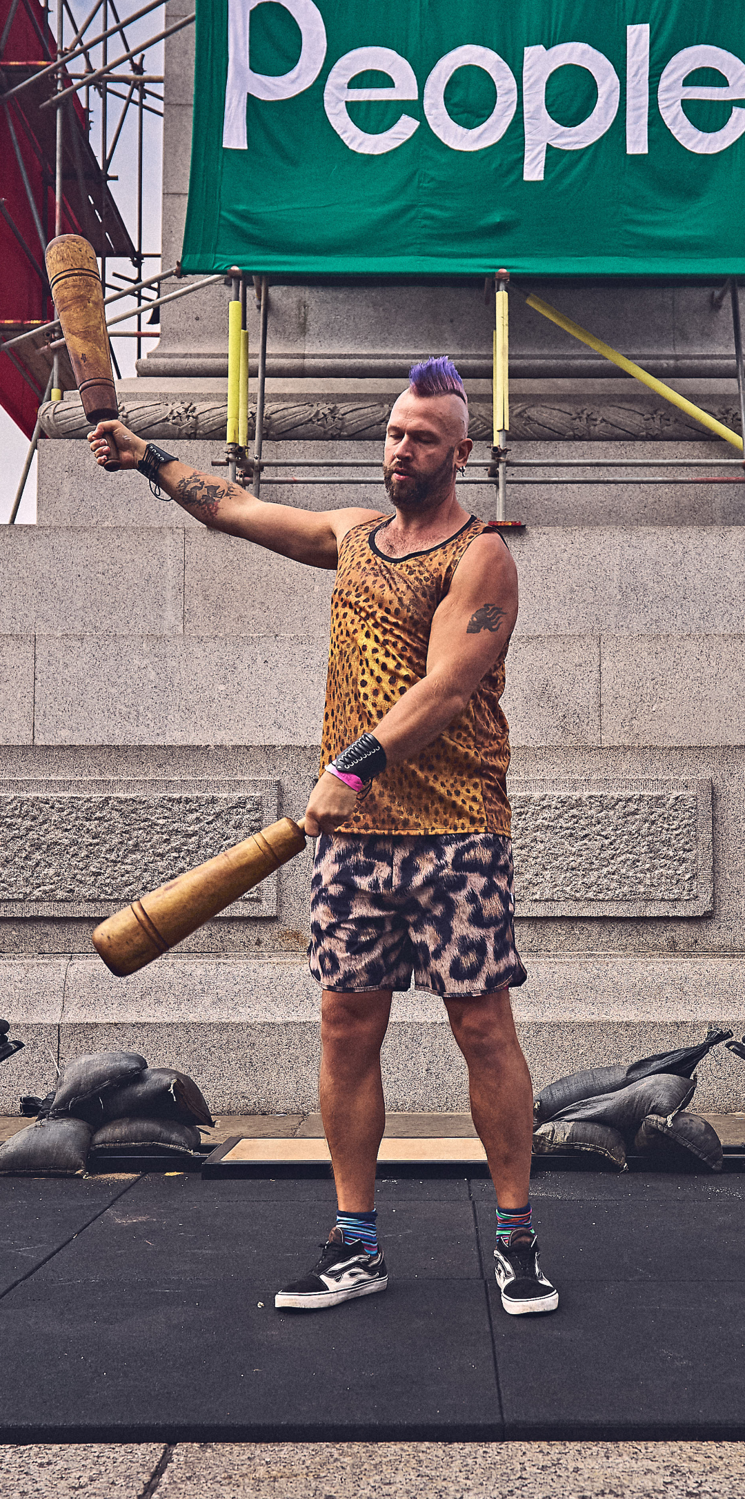 A man with purple hair and tattoos, wearing a leopard print tank top and shorts, swinging Persian meels in Trafalgar square London.