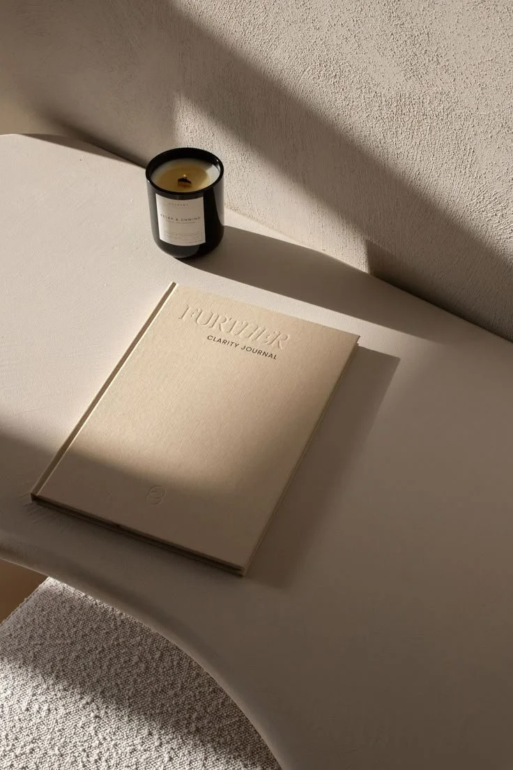 A beige journal labeled 'L’OURTIER CLARITY JOURNAL' closed on a white surface next to a black candle with cream-colored wax in a black jar. Sunlight casts shadows across the objects.
