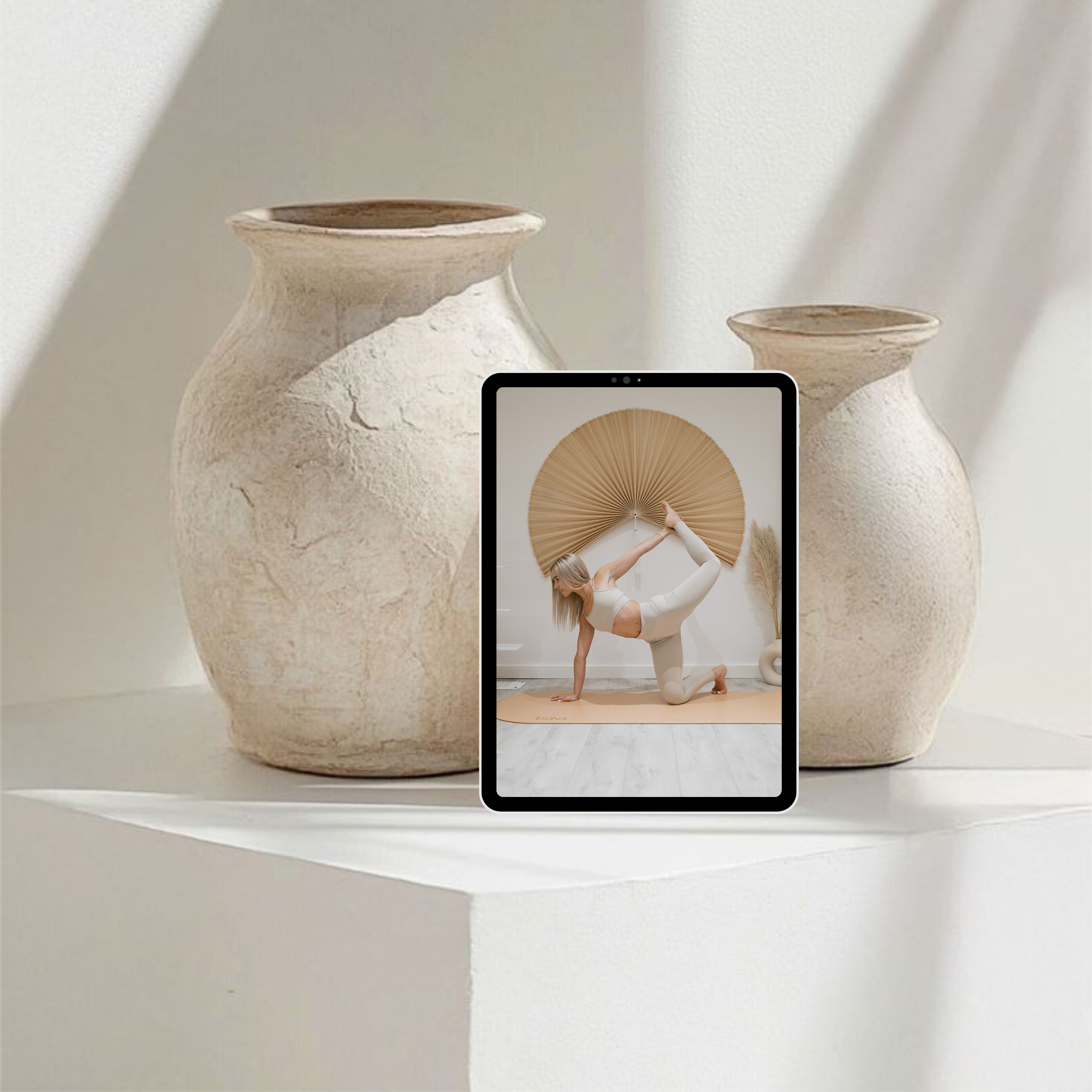 A tablet computer displaying a woman performing a yoga pose indoors, with a large decorative fan on the wall behind her, flanked by two vases, on a white surface.