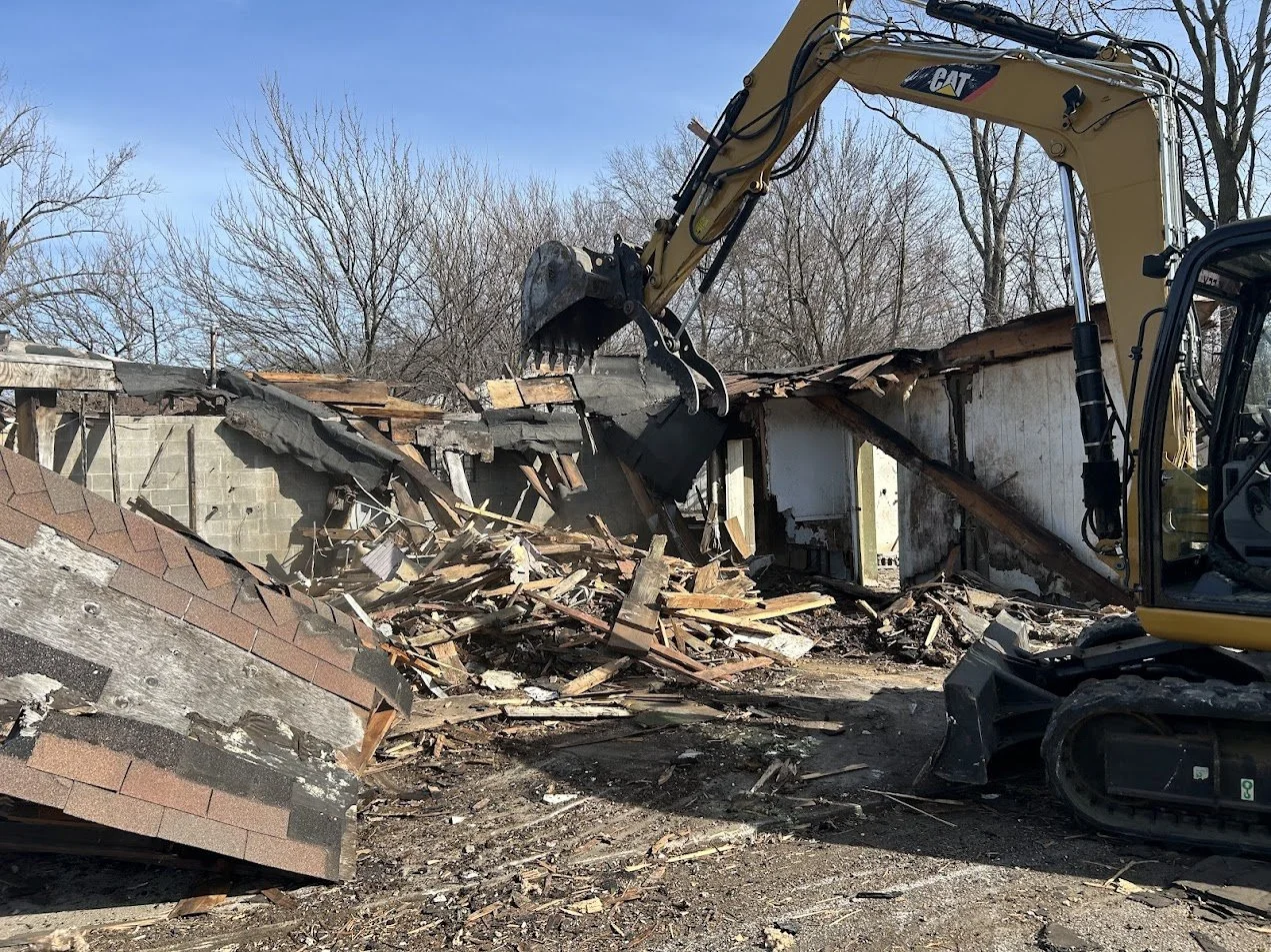 Excavator demolishing a building, debris and broken wood scattered around, clear sky in the background.