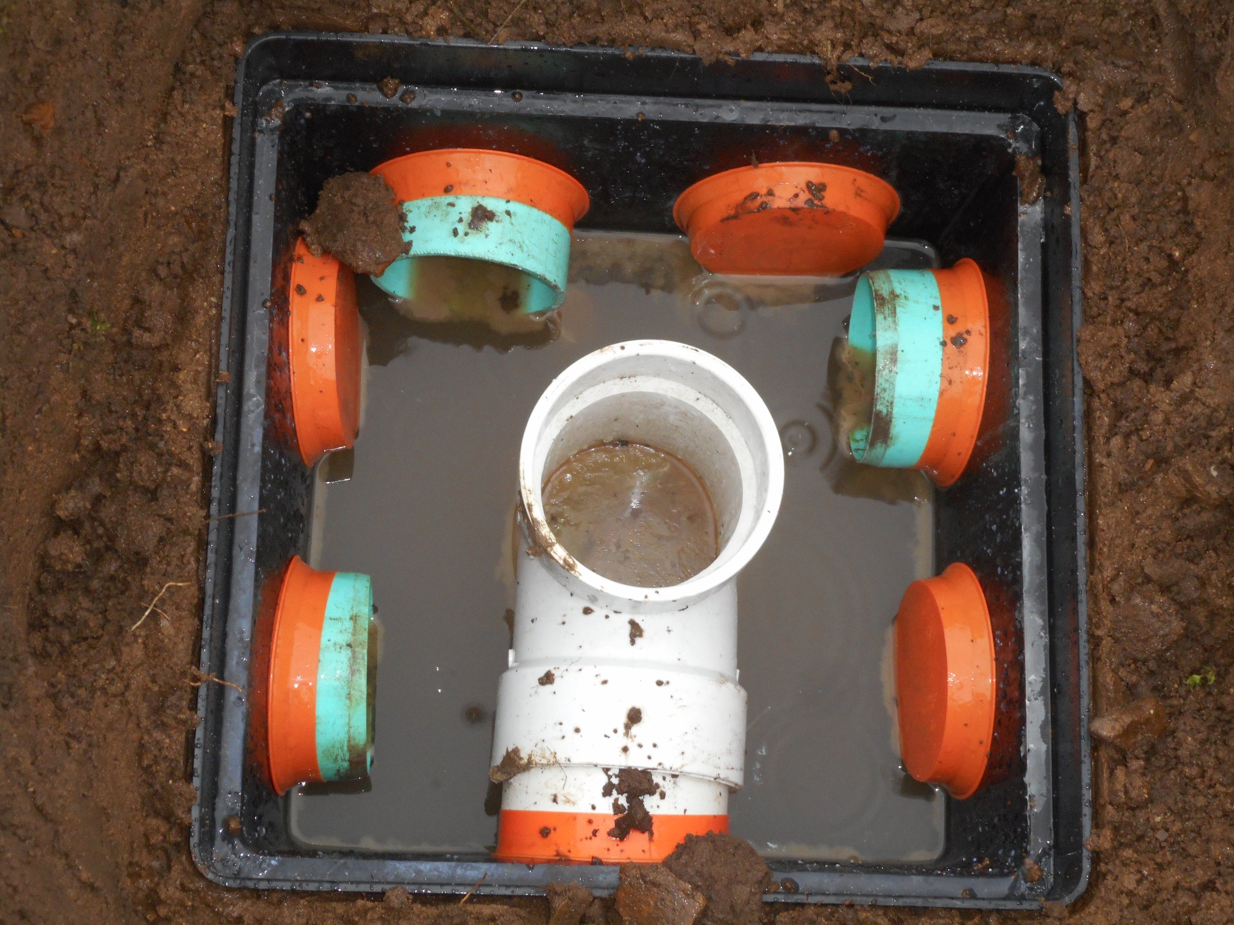 A square black plastic drainage system with a white pipe in the center, surrounded by five orange and teal PVC pipe connectors, all filled with dirty water and mud, set in dirt.
