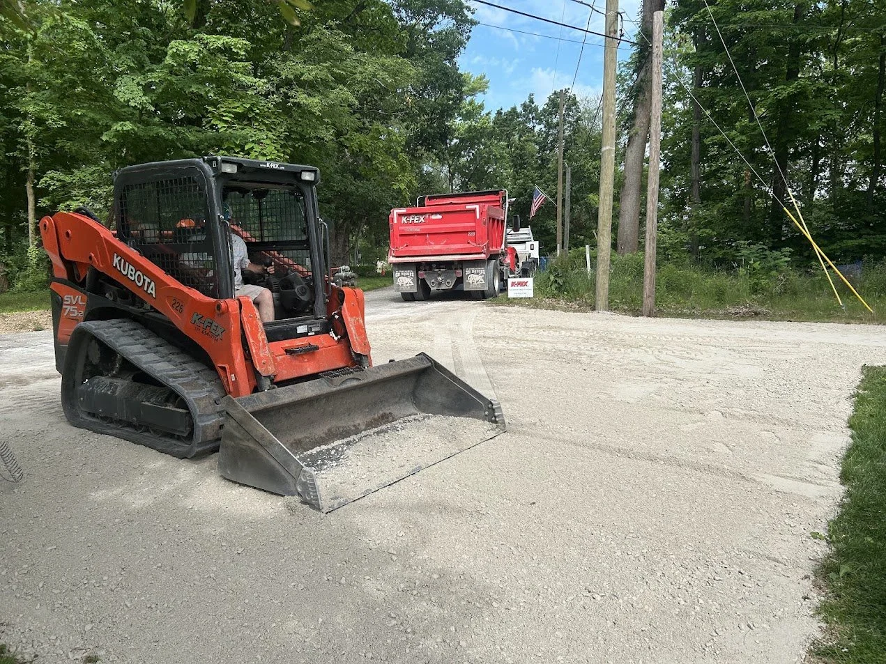 A K-FEX Kubota skidloader with a front bucket attachment is working on a gravel driveway in Saugany Lake