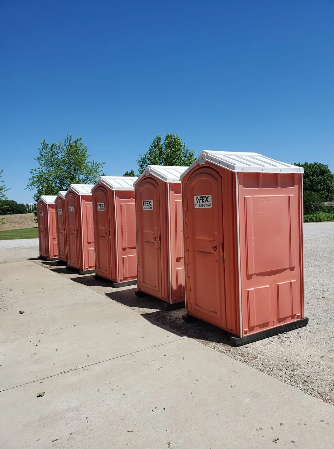 Row of five orange portable toilets outdoors on a sunny day with a clear blue sky and green trees in the background.