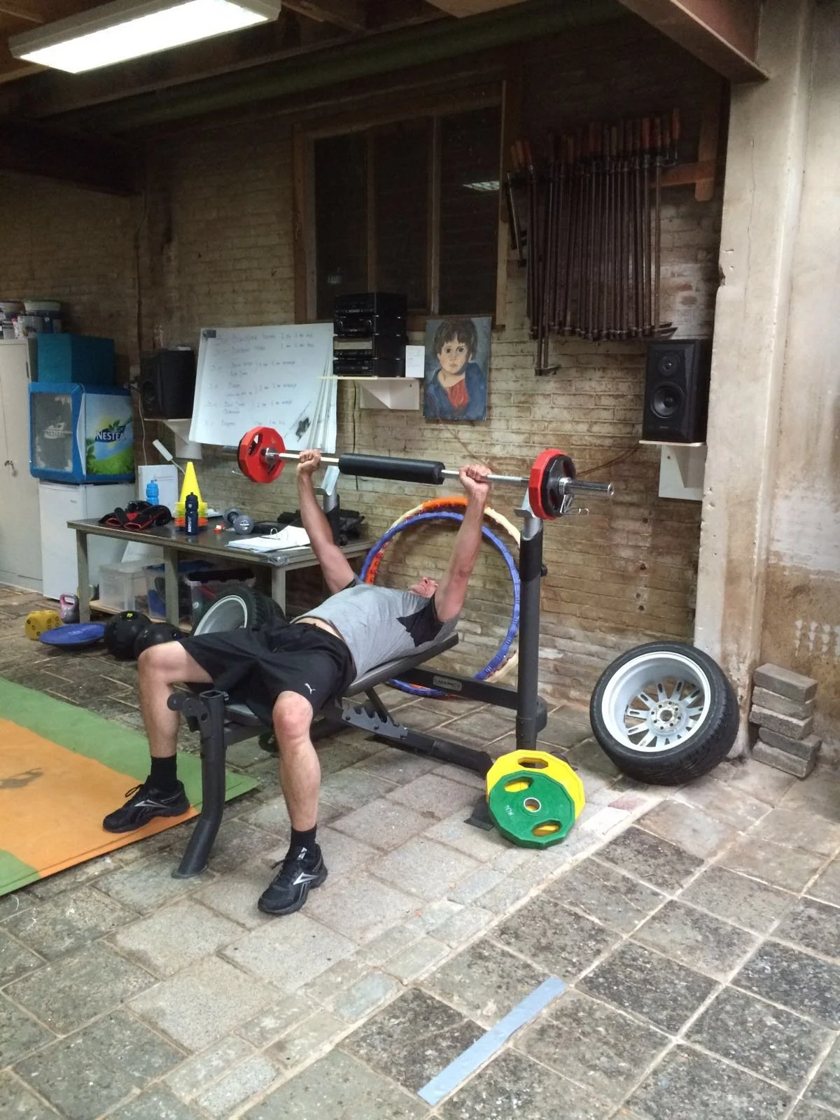 Persoon ligt op een fitnessbank en tilt een halter met rode gewichten in een sportschool of garage. Om de persoon heen liggen verschillende fitnessmaterialen en gereedschap, en op de muur hangen stangen en een portret van een kind.