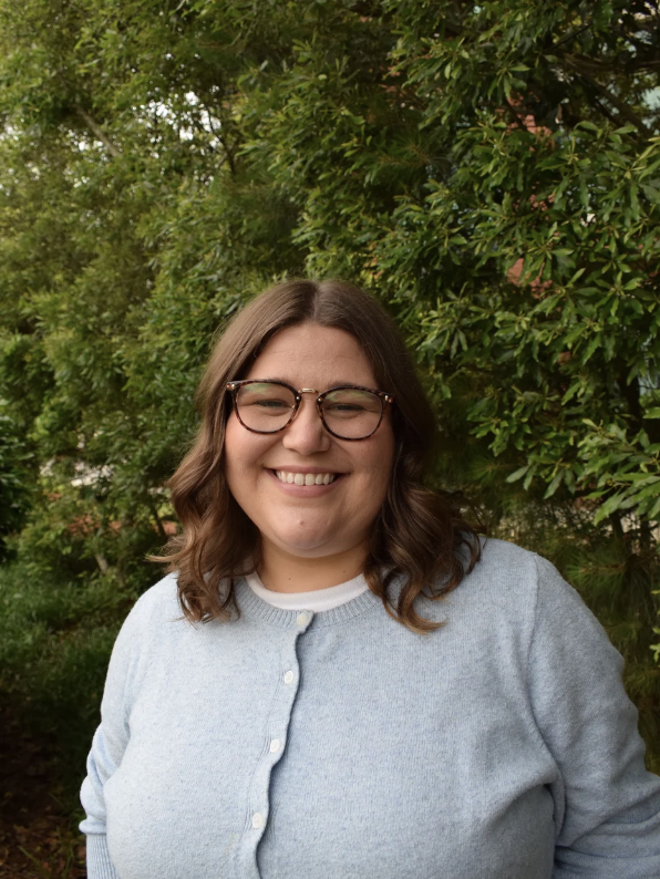 A woman with shoulder-length brown hair, glasses, and a light gray cardigan smiling outdoors in front of green foliage.