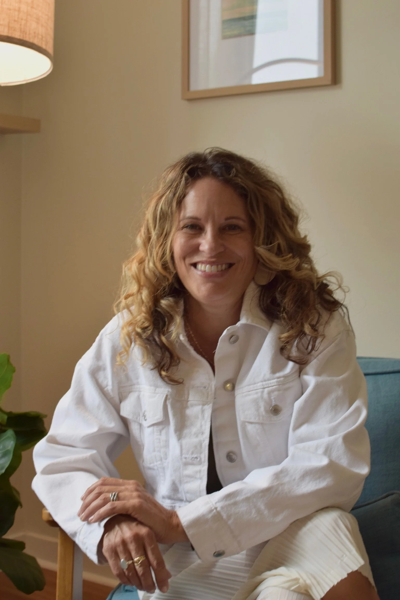 A smiling woman with curly brown hair wearing a white denim jacket, sitting on a chair in a room with beige walls, a blue couch, a framed picture, and a table lamp.