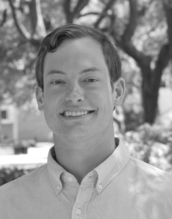 A young man with short dark hair smiling outdoors with a tree in the background.
