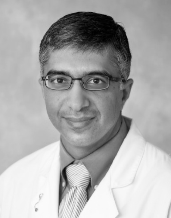 Black and white portrait of a man with short dark hair, glasses, and a slight smile, wearing a white lab coat and a striped tie.