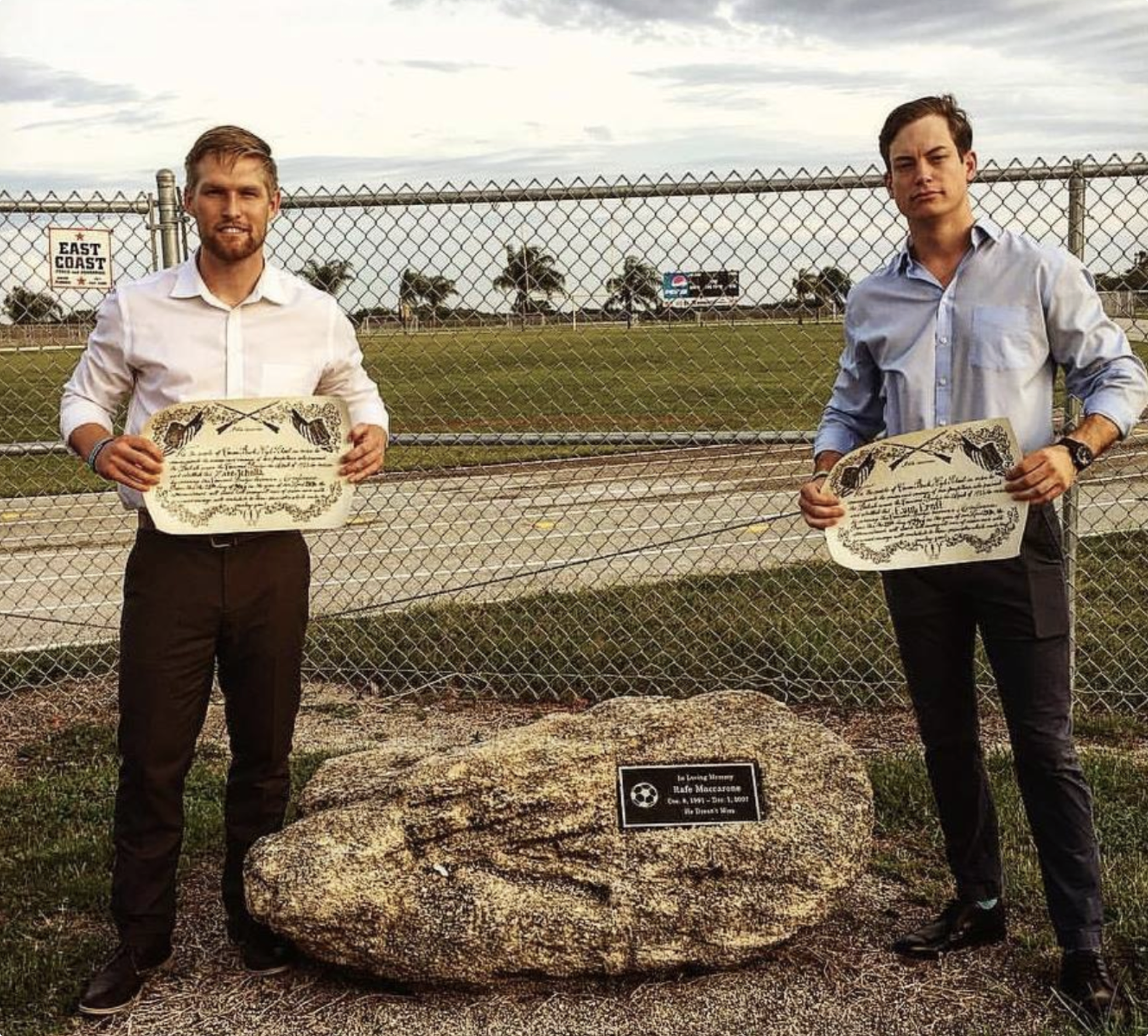 Two young men holding certificates, standing on either side of a memorial plaque for a boy named Rafe Maccarone, in front of a large rock with a neighborhood fence and grassy field in the background.