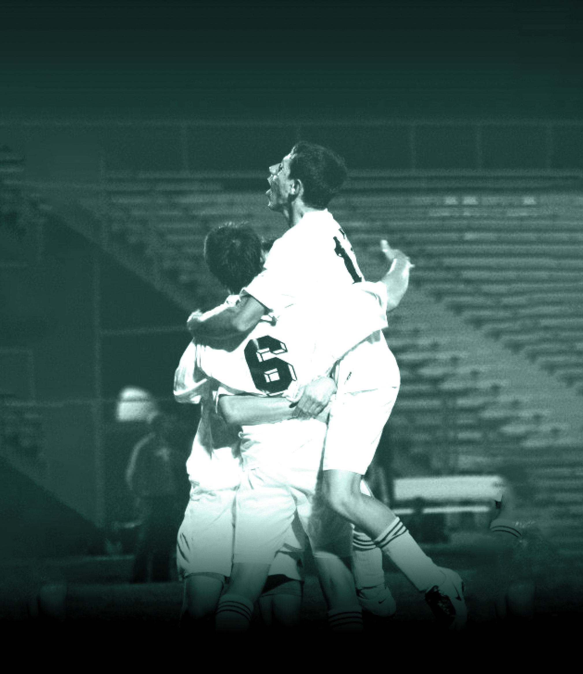 A group of young men celebrating on a soccer field, embracing each other after a goal or win, with a stadium in the background.