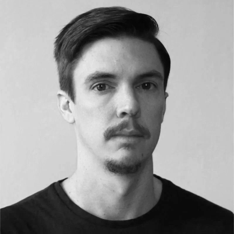 Black and white portrait of a young man with short, styled hair, slight facial hair, and a neutral expression, wearing a black shirt, standing against a plain light-colored wall.