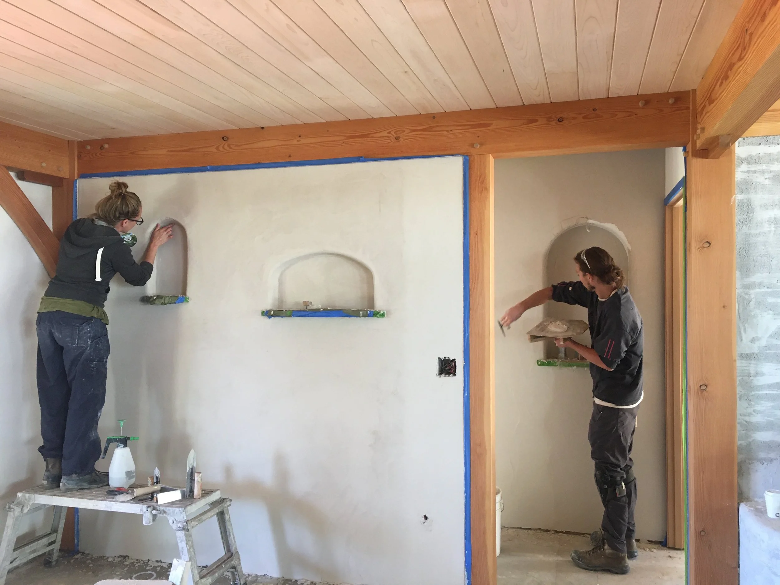 Two people working on wall renovation, one sanding drywall and the other with a trowel, in a room with wooden beams and no floor covering.