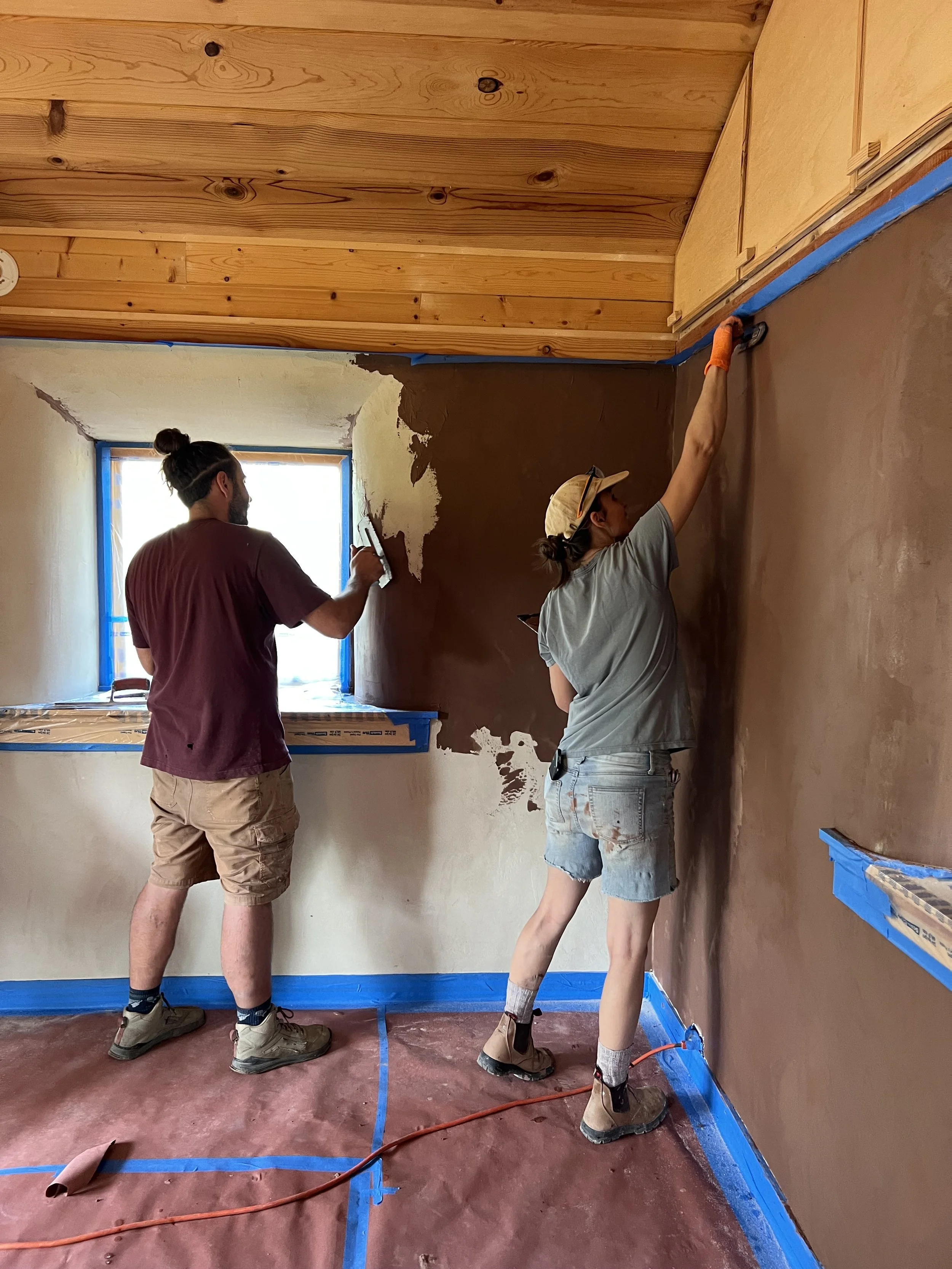 Two people are working on painting a wall inside a room. One person is standing near a window and holding a paintbrush, while the other person is reaching up to paint the upper part of the wall. The floor is covered with protective paper and painter's tape, and the ceiling is unfinished wood.