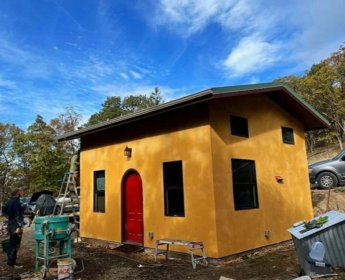 A small yellow stucco house with black-framed windows and a red door under a blue sky with some clouds, surrounded by trees, construction materials, and workers.