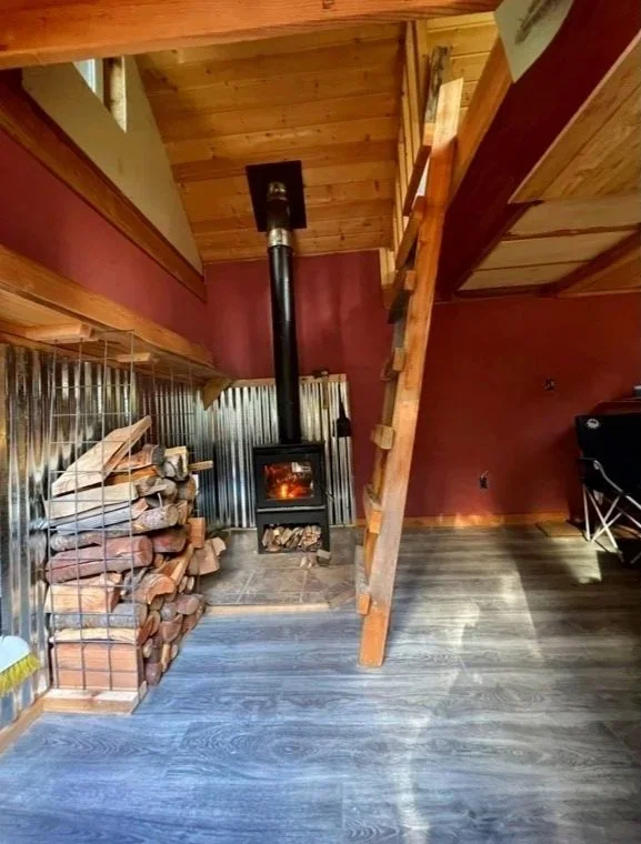 Wood stove with a fire burning, stacked firewood next to it, in a cozy wooden cabin interior.