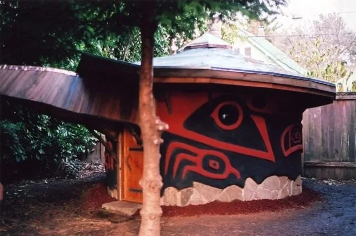 A small, round building with a roof that appears to be part of a traditional indigenous house, featuring black and red geometric and animal-like designs on its exterior walls, located outdoors surrounded by trees and a wooden fence.