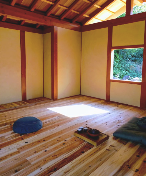 An empty traditional Japanese-style room with wooden floors, beige walls, a window showing greenery outside, a cushion, and a tray with teacups and a teapot.