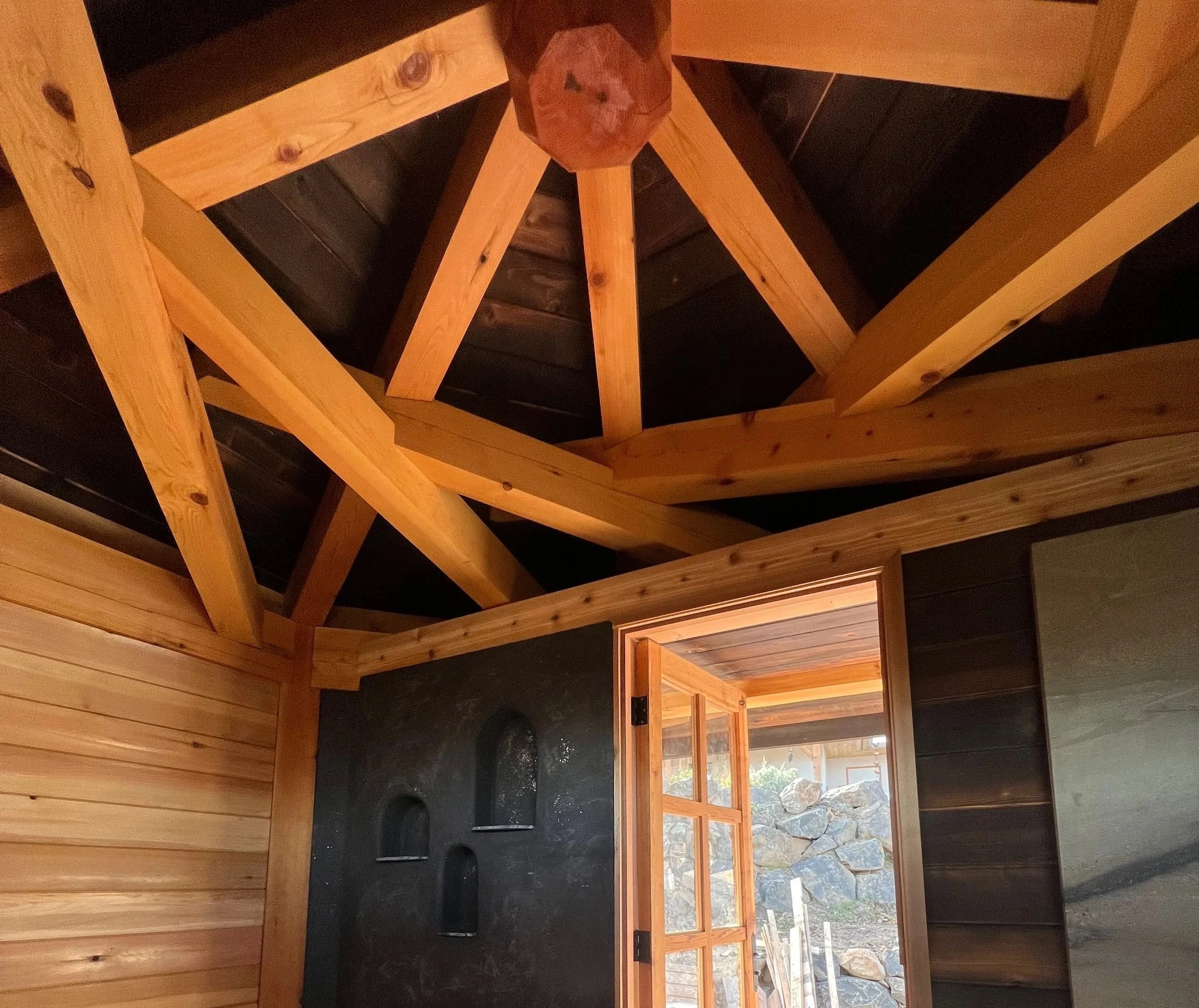 Interior view of a wooden structure under construction with visible beams, a window showing outdoor rocks, and a black surface with circular holes.