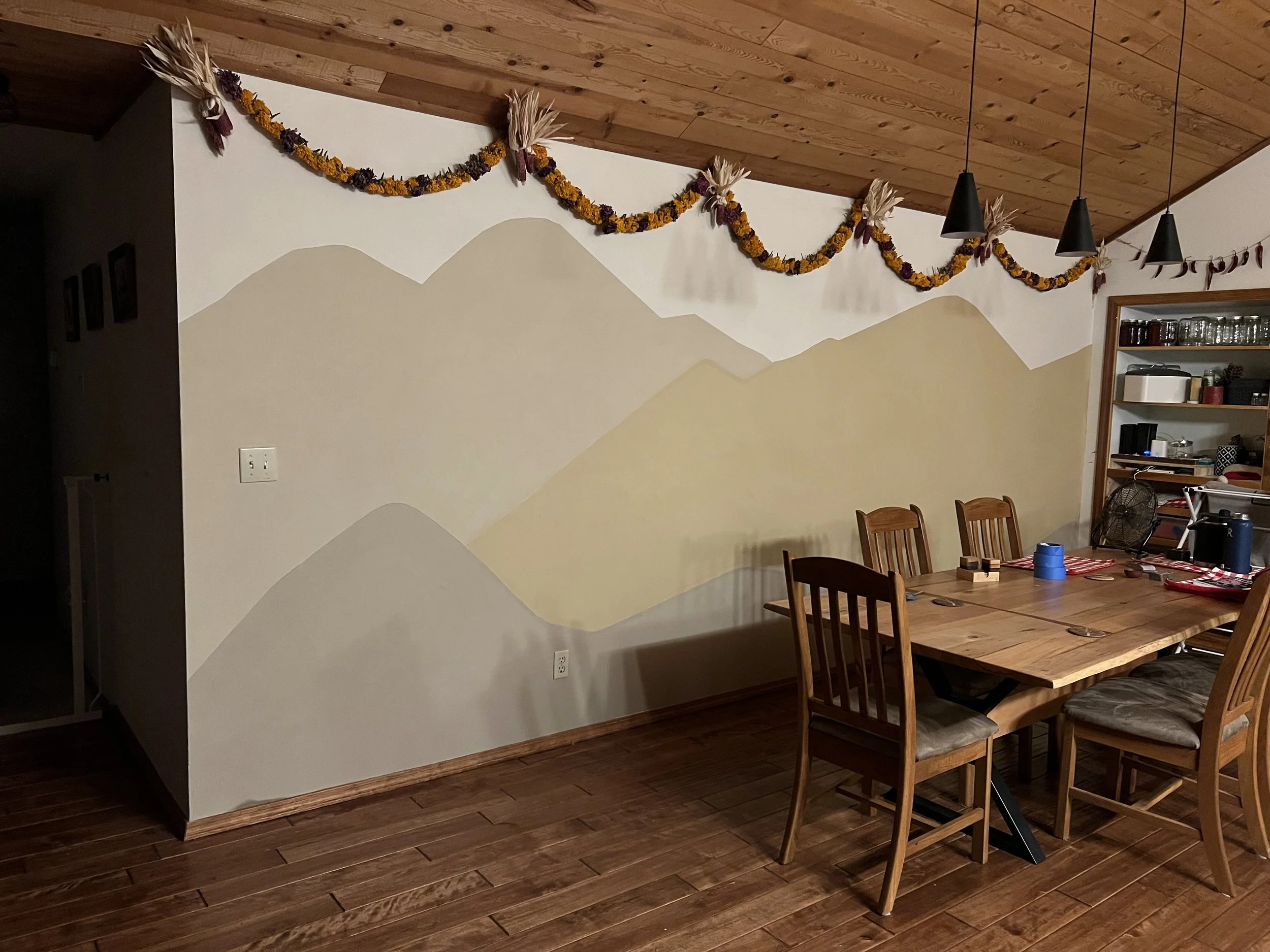 A wooden dining table with matching chairs in a room with a wood-paneled ceiling. A wall with a mountain landscape mural is decorated with a garland made of dried flowers and leaves. On the right side, there is a shelf with jars and kitchen items, and a partial view of a black fan.