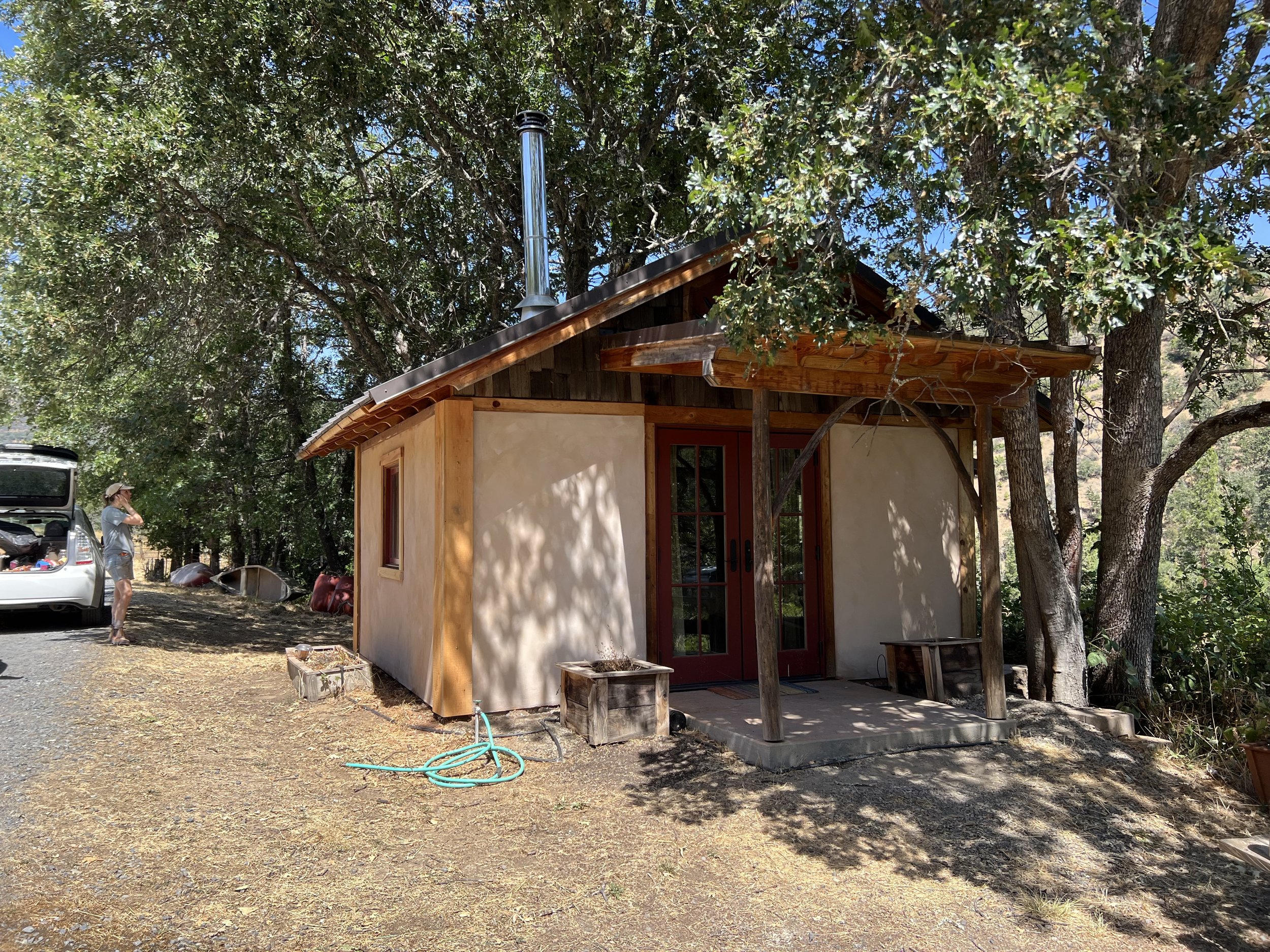 Small house with a wooden trimmed roof, red front door with glass windows, outdoors surrounded by trees with green leaves, sunlight creating shadows on the ground, a person standing near an open vehicle trunk, gardening or outdoor equipment present.