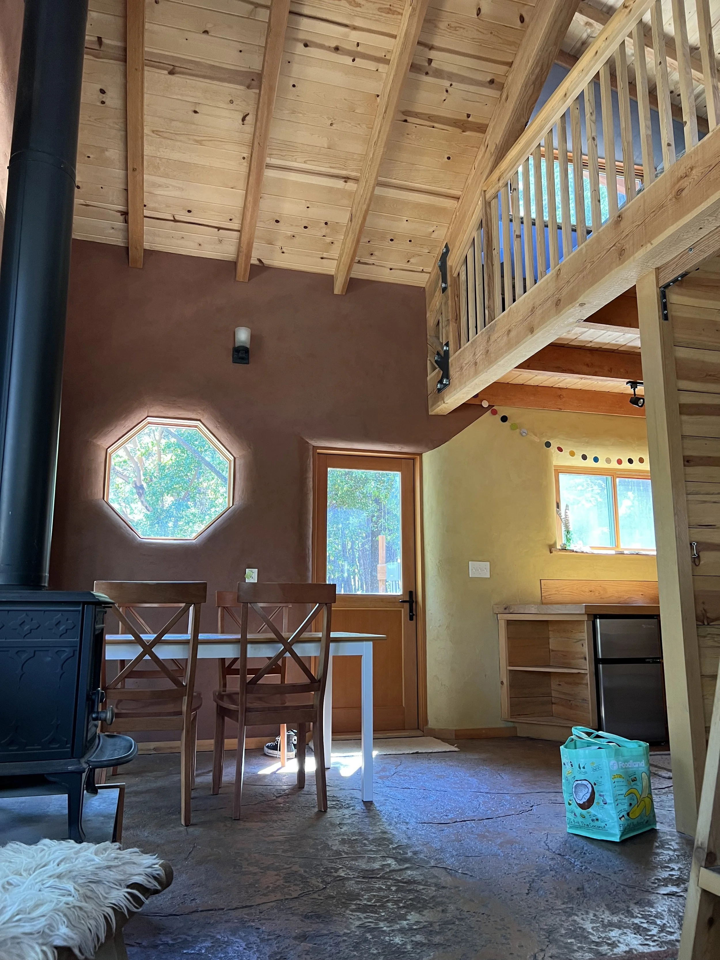 Interior view of a wooden cabin with a high ceiling, large windows showing trees outside, a wooden staircase leading to an upper level, a dining table with chairs, and a wood stove on the left side. There's a packet of food or snacks on the floor in the foreground.