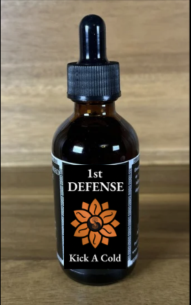 Small brown dropper bottle with a black label reading "1st DEFENSE" and "Kick A Cold," featuring an orange flower design, placed on a wooden surface.