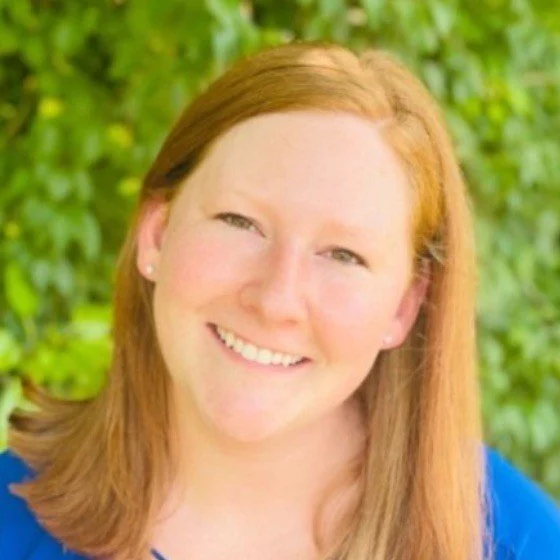 A smiling woman with red hair outdoors with green foliage in the background.