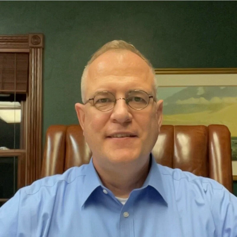 A man with glasses and short blond hair, wearing a light blue dress shirt, sitting in a brown leather chair in front of a green wall and a framed landscape painting.