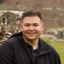 A man smiling outdoors, wearing a black jacket with a blurred natural background.