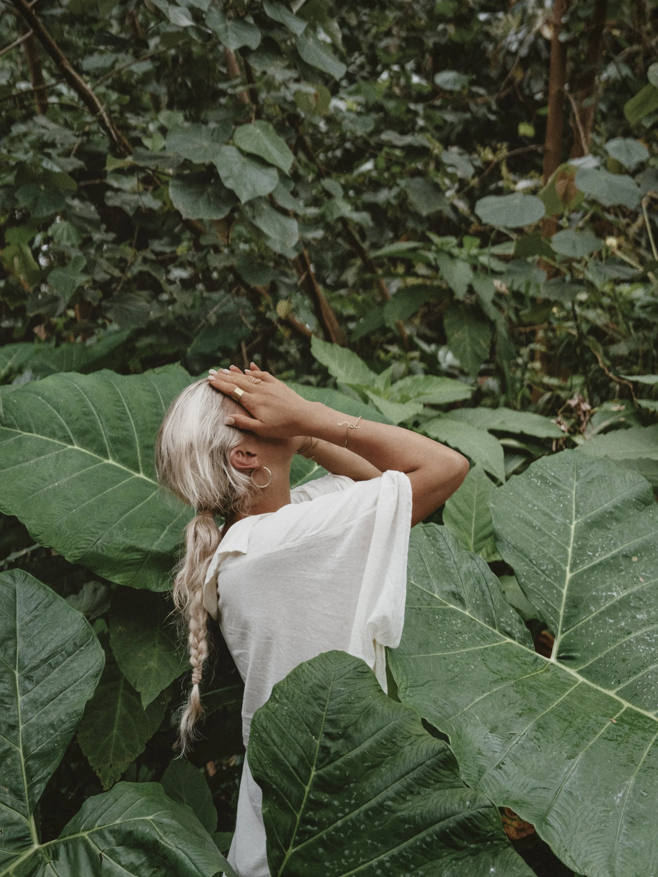 A woman with blonde hair in a braid, wearing a white shirt, is standing among large green leaves and touching her head with her left hand, surrounded by dense foliage.