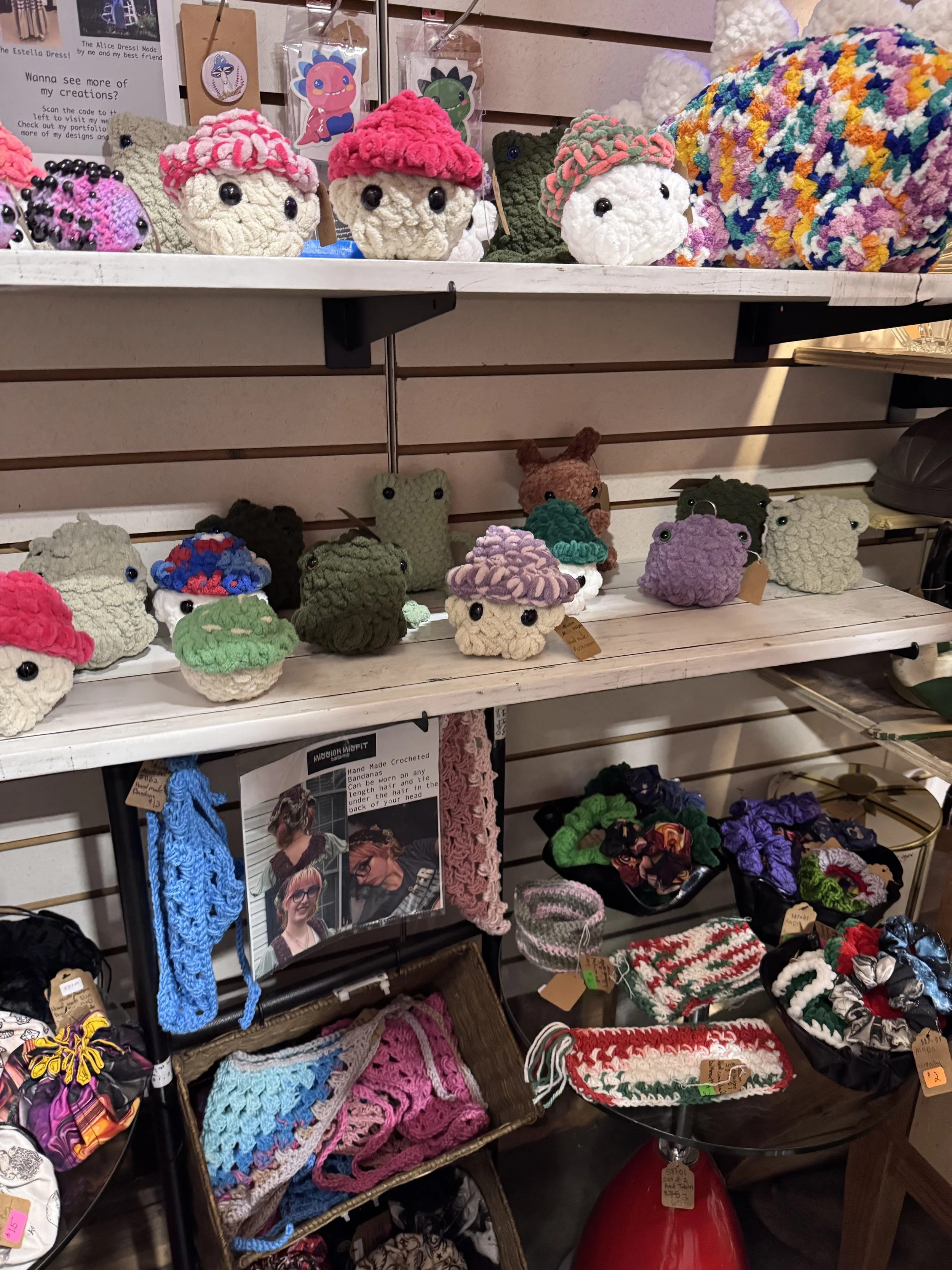 Crocheted plush toys resembling cupcakes, frogs, and other animals displayed on shelves. Various colorful crochet accessories and craft items on table and hanging on wall.