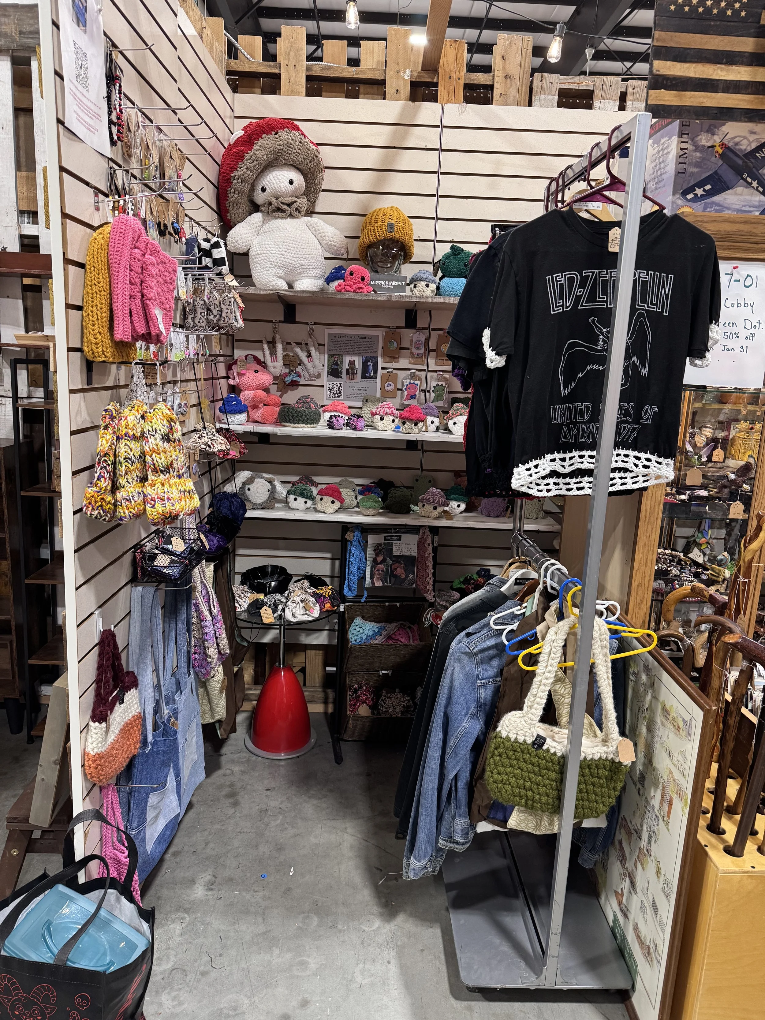 A display of handmade crochet dolls, hats, and bags at a store, including a large plush doll with a mushroom hat, assorted small crocheted animals, and clothing items like a Led Zeppelin T-shirt and denim jackets.