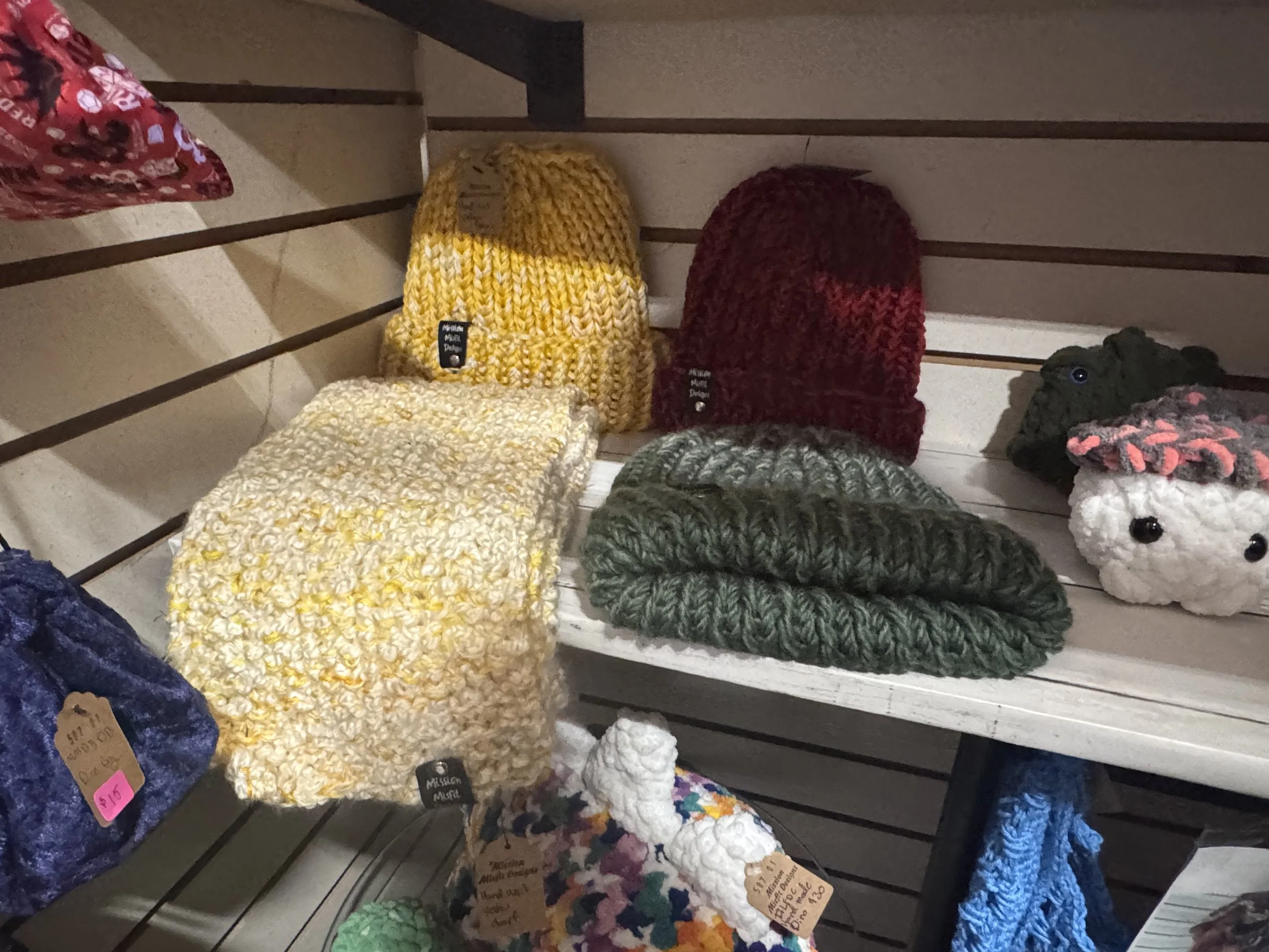 Display of handmade knitted and crocheted hats, scarves, and a plush sheep on a slatted shelf.