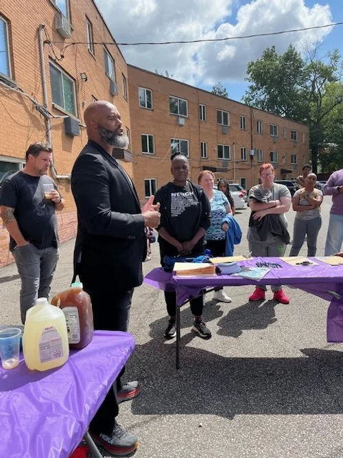 A man in a black suit speaking to a group of people outdoors in an alleyway with apartment buildings in the background, with tables covered in purple cloth and food condiments.