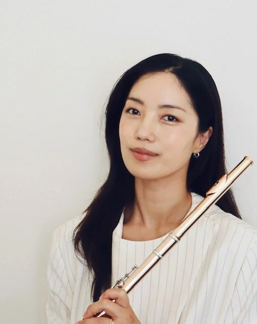 A young woman with long dark hair holding a flute, standing against a plain white background.