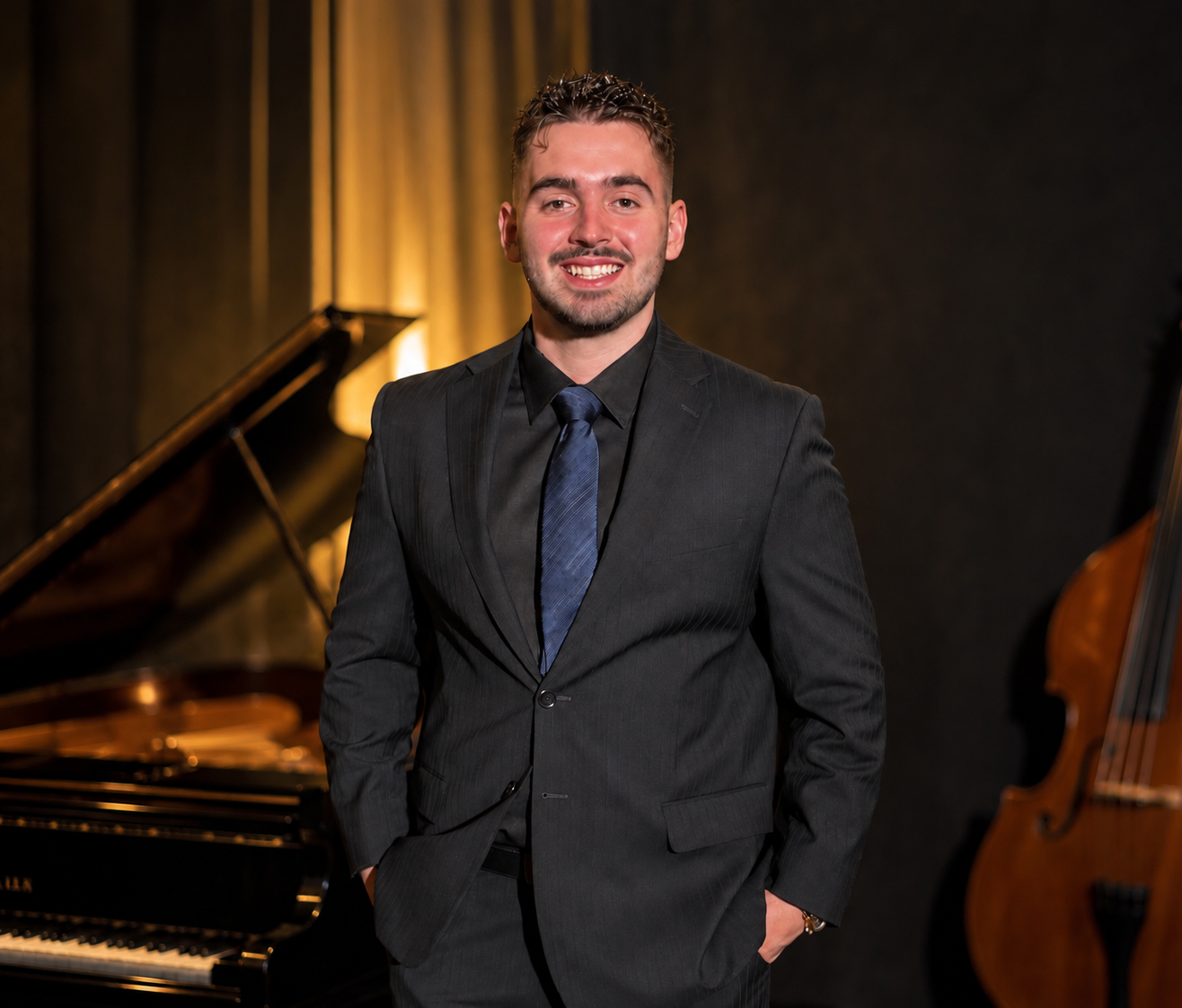 Young man in a dark suit and blue tie standing in a music rehearsal room with a piano and bass guitar in the background.