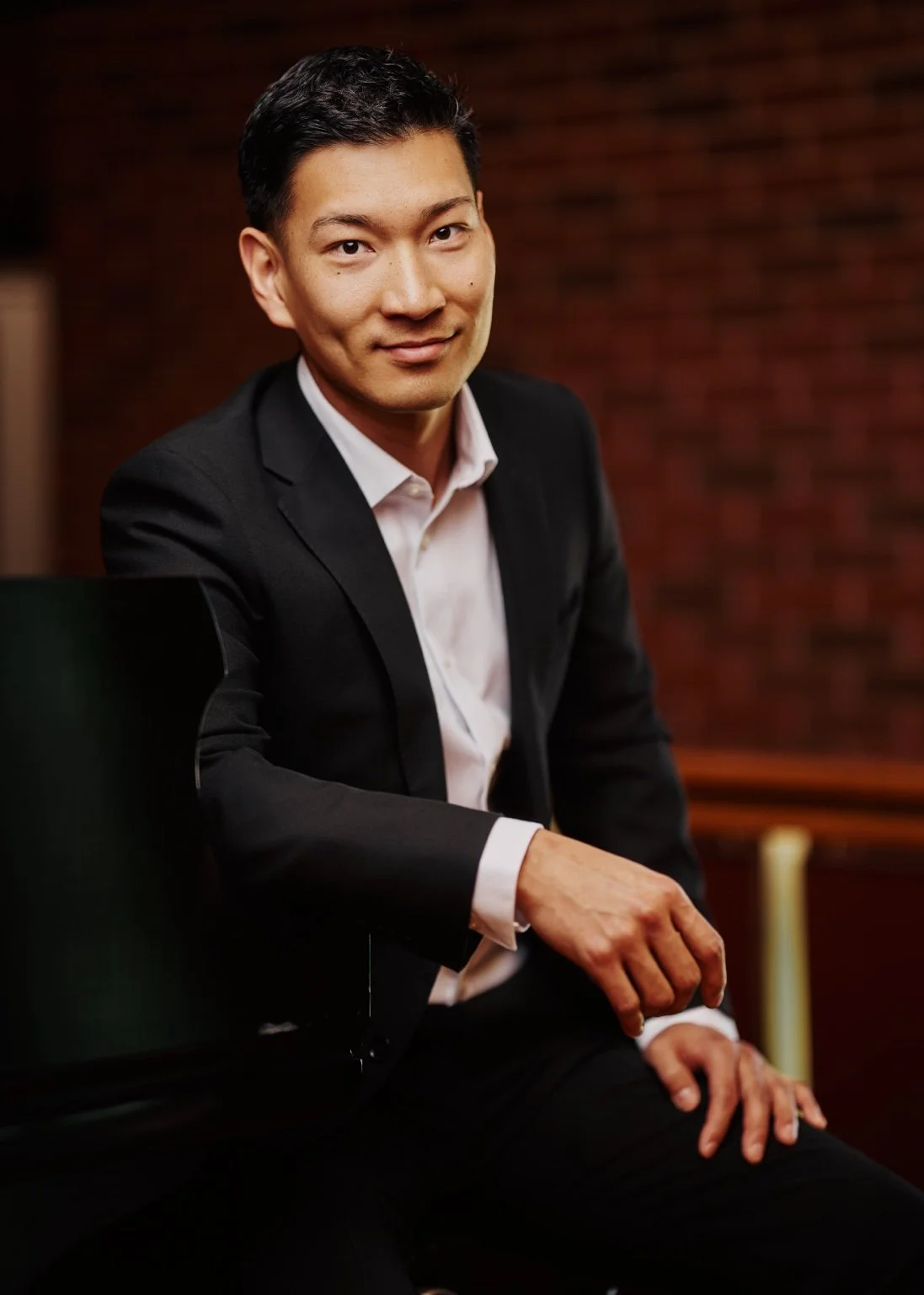 A man in a black suit sitting at a piano with a red brick wall in the background.